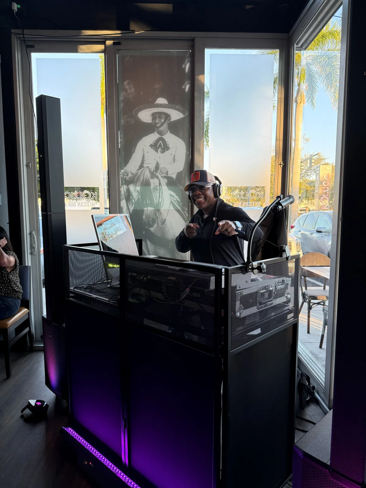 ShaCoda pointing to the camera from behind his DJ booth, with palm trees outside the windows